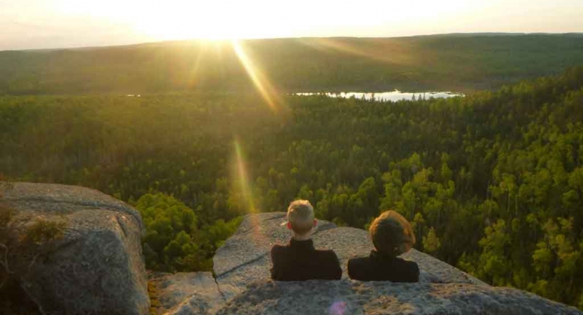 two students rest against rocks overlooking a vast green forest containing a body of water